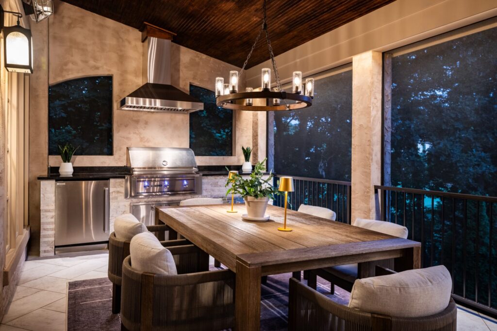 Covered porch with outdoor kitchen and dining area featuring a wood ceiling, stainless steel grill, and modern chandelier lighting.